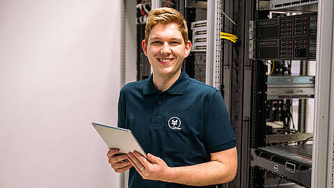 IT-Systemelektroniker (m/w/d) Auszubildender zum IT-Systemelektroniker (m/w/d) steht vor einem Serverschrank und hält ein Tablet in der Hand