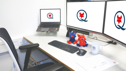 Moderner Arbeitsplatz beim Q-FOX® Höhenverstellbarer, weißer Schreibtisch mit Laptop und zwei großen Bildschirmen, auf denen das Q-FOX® Logo zu sehen ist. Vor den Bildschirmen sitzen ein blauer und ein roter Q-FOX®, außerdem sind auf dem Schreibtisch eine Q-FOX® Tasse, ein Block mit Kugelschreiber sowie eine Tastatur und Maus zu sehen.