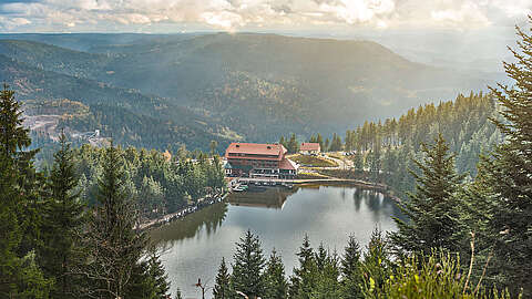 Regionaler Arbeitgeber im Schwarzwald Eine große Hütte mit rotem Dach liegt an einem kleinen See, umgeben von Kiefern und bewaldeten Hügeln unter einem wolkenverhangenen Himmel, durch den die Sonnenstrahlen scheinen.