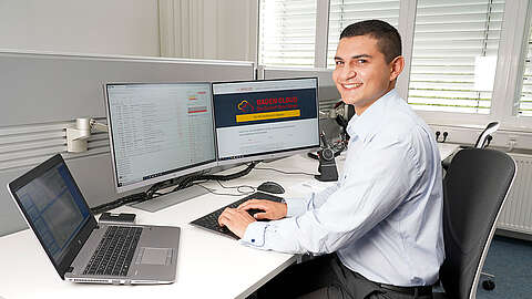 Trainee beim Q-FOX® Junger Mitarbeiter sitzt an einem PC mit mehreren Bildschirmen und tippt in die Tastatur