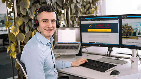 Einstiegsmöglichkeiten für Schüler beim Q-FOX® Q-FOX® Mitarbeiter sitzt an einem Schreibtisch mit PC-Monitoren und hat ein Headset auf
