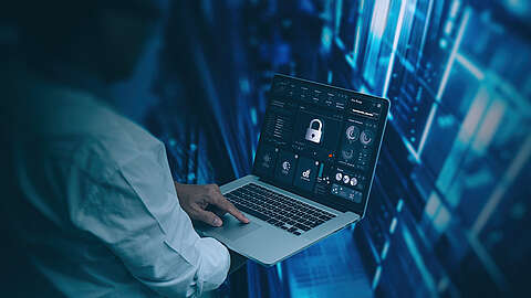 Serverraum Eine Person, die einen Laptop benutzt, auf dem Symbole für Cybersicherheit und Datenschutz mit einem Vorhängeschloss-Symbol auf dem Bildschirm angezeigt werden, steht vor einem digitalen blauen Hintergrund, der Server oder Datennetze darstellt.