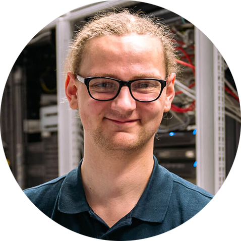 Luk Jansen, Auszubildender zum Fachinformatiker für Systemintegration bei der LEITWERK AG Eine Person mit heller Haut, Brille und zurückgebundenem, langem, hellem Haar, lächelnd und in einem dunklen Hemd mit Kragen steht vor Computerservern, im Hintergrund sind Kabel und Geräte sichtbar.